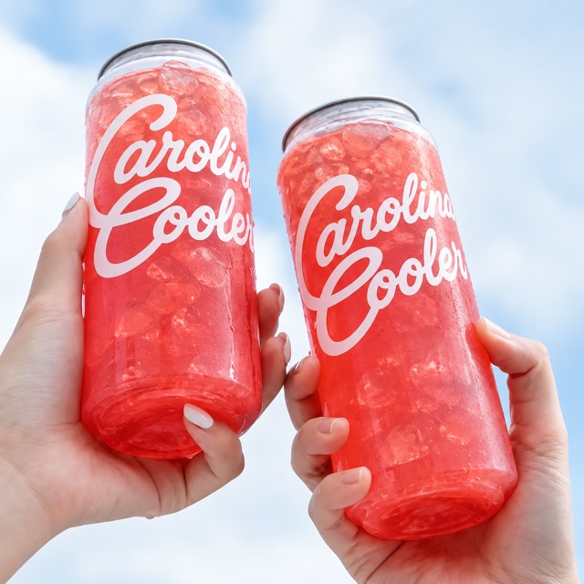 Two cans of red Carolina Cooler fruit tea raised against a summer sky